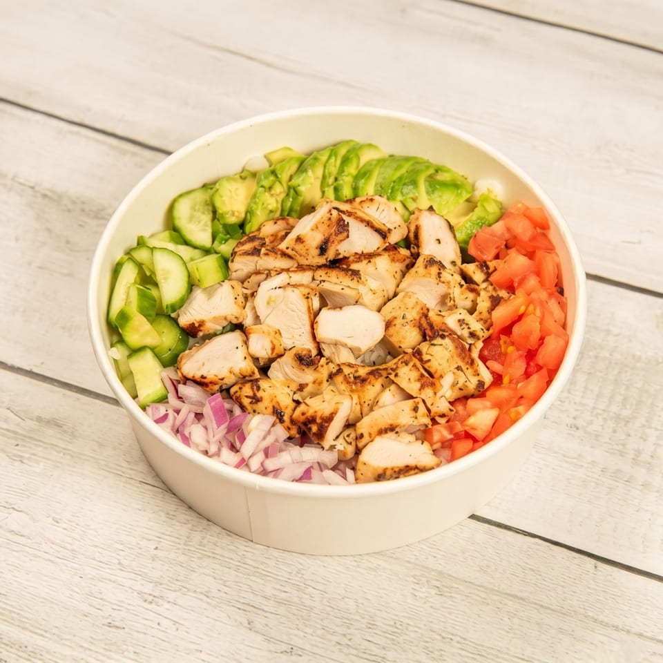 Grilled chicken, avocado, cucumber, tomatoes, and red onions in a white bowl on a wooden surface.