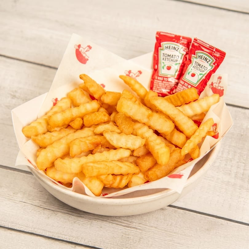 A white bowl filled with golden crinkle-cut fries and two Heinz ketchup packets on a wooden table.